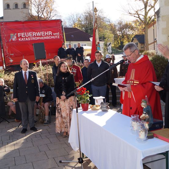 Eine religiöse Zeremonie findet im Freien statt. Ein Mann in einem roten Gewand steht an einem Rednerpult und liest ein Buch. Hinter ihm steht ein Tisch, der mit einem weißen Tuch bedeckt ist und verschiedene Gegenstände darauf hat. Menschen stehen hinter dem Tisch und eine Frau ist im Vordergrund.