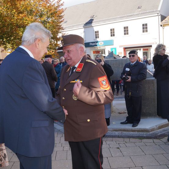 Zwei Männer in Uniformen schütteln sich die Hände auf einem Freigelände. Hinter ihnen stehen mehrere Personen und beobachten. Der Ort ist von einem Ziegelboden und einem großen Pfeiler umgeben. Der Himmel ist klar.