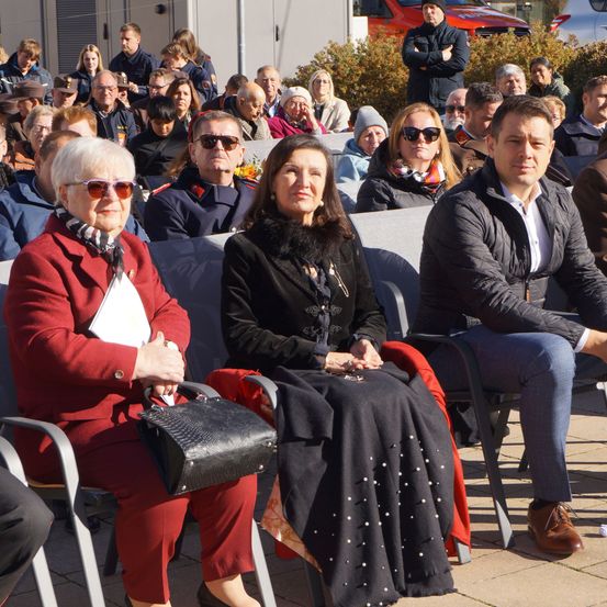 Eine Outdoor-Veranstaltung mit einer Menschenmenge. Mehrere Frauen und Männer sitzen auf Stühlen, darunter eine Frau mit einer Handtasche und eine andere Frau mit einem Schal. Ein Mann sitzt ebenfalls und schaut nach rechts.