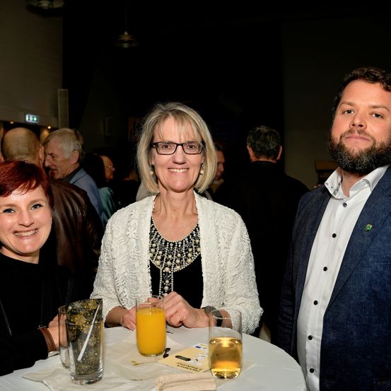 Drei Menschen lächeln in die Kamera. Sie sitzen um einen Tisch mit Getränken herum. Sie sind bei einer gesellschaftlichen Veranstaltung. Der Raum ist schwach beleuchtet.