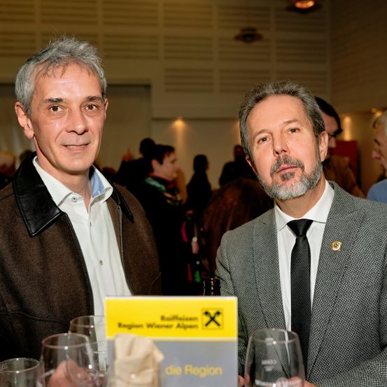 Zwei Männer sitzen an einem Tisch in einem überfüllten Raum, einer trägt eine Jacke und der andere einen Anzug. Sie haben ein gelbes Menü vor sich und scheinen zu unterhalten.