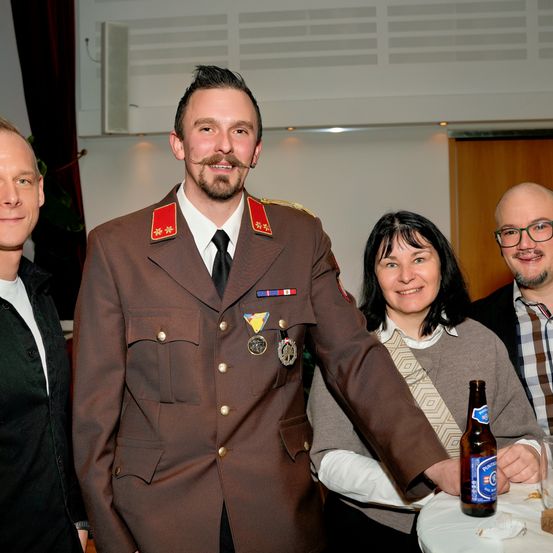 Vier Personen versammeln sich zu einem Foto, ein Mann in Uniform mit Medaillen, eine Frau mit Brille und zwei andere Männer, einer in einem schwarzen Jackett und der andere in einem karierten Hemd, mit einer Bierflasche auf einem Tisch.