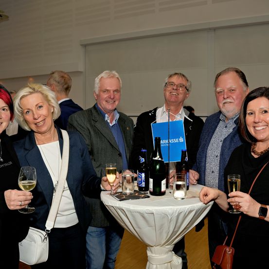 Eine Gruppe von Menschen steht um einen Tisch mit Weingläsern und Flaschen. Eine Person hält ein blaues Arkasse-Schild. Die Gruppe scheint sich bei einer gesellschaftlichen Veranstaltung zu befinden.