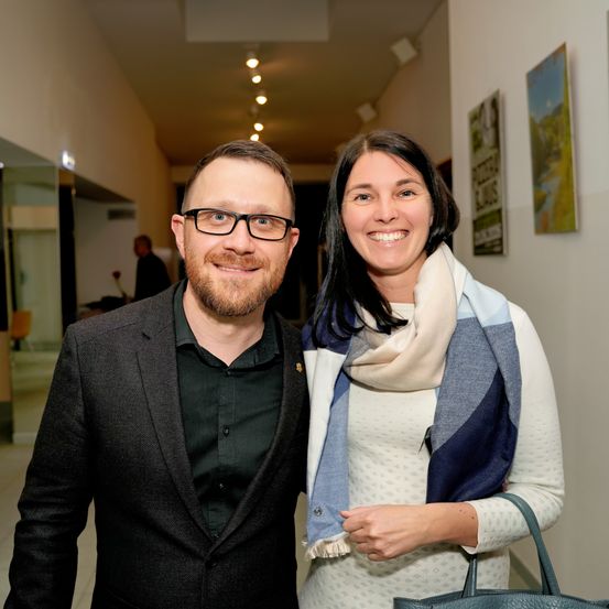 Ein Mann und eine Frau stehen lächelnd in einem Flur. Der Mann trägt einen schwarzen Anzug und eine Brille, während die Frau einen weißen Pullover und einen blau-weißen Schal trägt. Sie posieren für ein Foto.