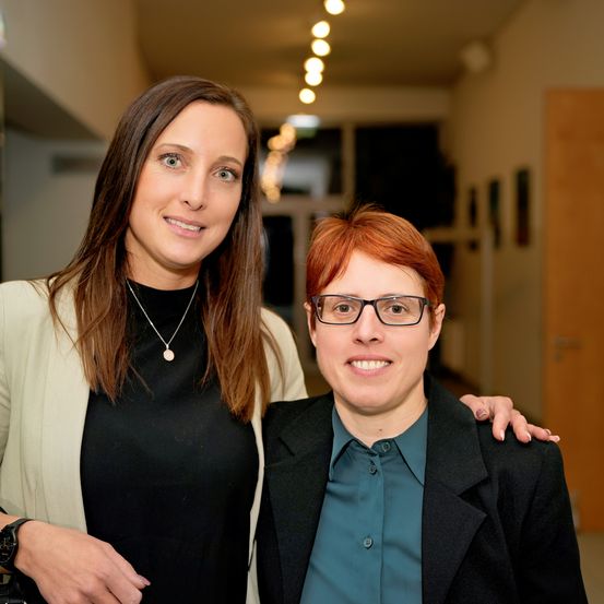Zwei Frauen stehen eng beieinander und lächeln in einem gut beleuchteten Korridor. Die Frau auf der linken Seite trägt eine Halskette und eine beigefarbene Strickjacke. Die Frau auf der rechten Seite hat eine Brille und einen schwarzen Blazer an.