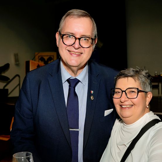 Ein Mann und eine Frau lächeln und posieren für ein Foto. Der Mann trägt eine Brille, einen Anzug und eine Krawatte. Die Frau trägt ein weißes Rollkragenshirt und eine Brille. Sie stehen eng beieinander. Im Hintergrund ist eine verschwommene Treppe und eine Wand zu sehen.