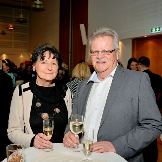 Ein Mann und eine Frau sitzen an einem Tisch, lächeln und halten Gläser mit Wein. Dahinter steht eine Gruppe von Menschen in einem Raum mit einer Bühne und einer Tür.