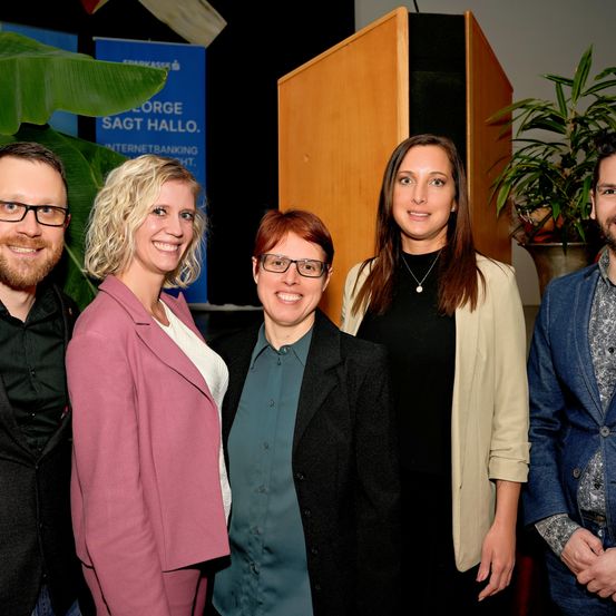 Fünf Menschen lächeln und posieren für ein Foto. Zwei Frauen und zwei Männer tragen Brillen. Eine Frau hat lange Haare und trägt eine Halskette. Blumentöpfe stehen im Hintergrund.