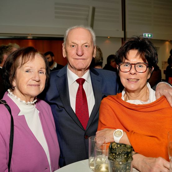 Drei ältere Personen lächeln und posieren für ein Foto bei einer Veranstaltung, der Mann in Anzug und roter Krawatte und zwei Frauen in farbenfroher Kleidung.