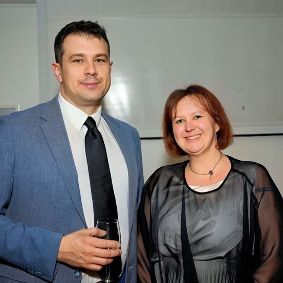 Ein Mann und eine Frau stehen nah beieinander und lächeln in die Kamera. Der Mann hält ein Weinglas. Beide sind formell gekleidet.