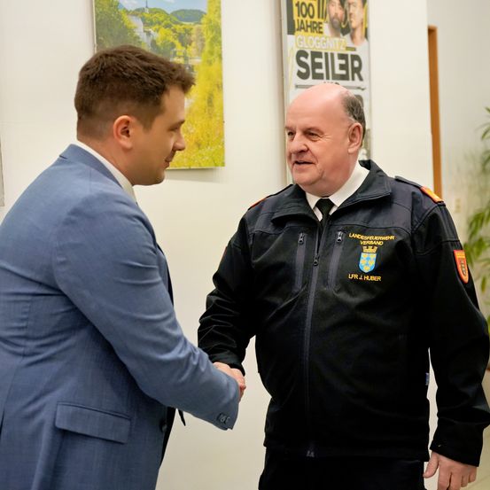 Zwei Männer, einer im Anzug und der andere in einer Polizeiuniform, schütteln sich die Hände in einem Büro.