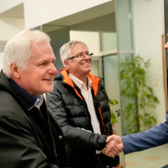 Drei Männer in einem Flur, zwei lächeln und schütteln Hände, einer trägt eine Brille und eine orange Jacke.