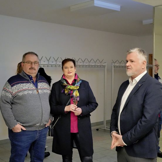 Drei Menschen stehen in einem Raum. Der Mann auf der linken Seite trägt eine Brille und einen grauen Pullover. Die Frau in der Mitte trägt ein rosa Hemd und einen Schal. Der Mann auf der rechten Seite trägt einen Anzug und hält ein Glas. Dahinter hängen einige Kleidungsstücke an den Kleiderbügeln.