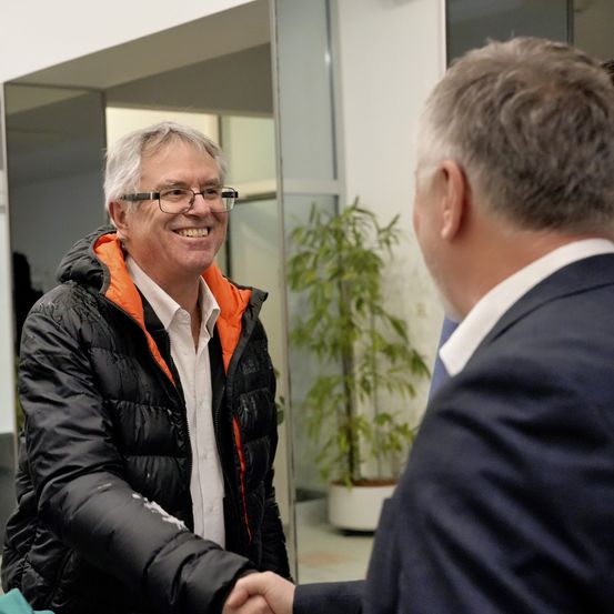 Ein Mann mit Brille und einer schwarzen und orangefarbenen Jacke lächelt, während er einem anderen Mann im Anzug die Hand schüttelt, in einem Gebäude mit einer Pflanze im Hintergrund.