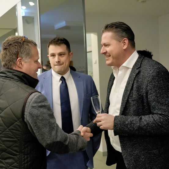Drei Männer in formeller Kleidung unterhalten sich. Der Mann auf der linken Seite trägt eine schwarze Jacke und hat eine Brille auf dem Kopf. Der Mann in der Mitte hält ein Glas Wein und trägt einen blauen Anzug mit Krawatte. Der Mann auf der rechten Seite lächelt und hält ebenfalls ein Glas.
