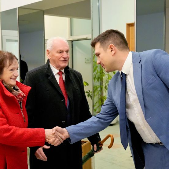 Drei Personen lächeln in einem modernen Gebäude. Eine Frau in einem roten Mantel schüttelt die Hand eines Mannes in einem blauen Anzug. Ein älterer Mann steht hinter ihnen.