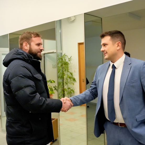 Zwei Männer schütteln sich in einem Büro die Hände. Einer trägt einen blauen Anzug, der andere eine schwarze Daunenjacke. Hinter ihnen ist die Reflexion einer Pflanze und einer Holztür zu sehen.