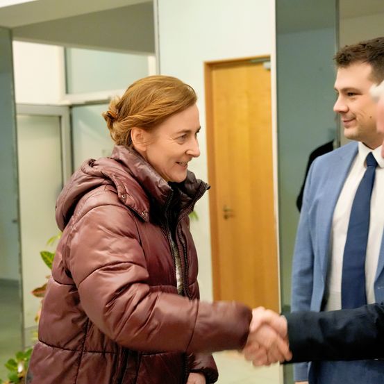 Eine Frau in einer braunen Daunenjacke schüttelt die Hände zweier Männer, einer in einem blauen Anzug und der andere in einem schwarzen Anzug. Die Szene spielt sich drinnen ab, in der Nähe einer Glaswand.