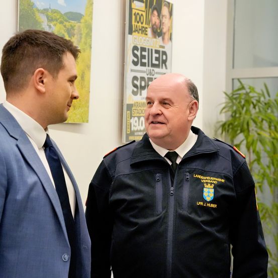 Zwei Männer stehen in einem Raum, einer in einer Marinejacke mit Logo, der andere in einem blauen Anzug. Hinter ihnen ein Plakat für '100 Jahre Gloggnitzer' und eine grüne Pflanze.