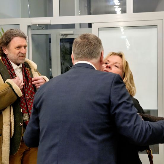 Drei Personen stehen in einem Raum in der Nähe einer Glastür. Ein Mann im Anzug beugt sich vor zu einer Frau, während ein Mann mit einem Schal sie ansieht.