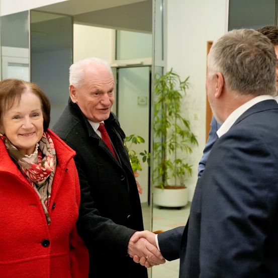 Ein älterer Mann und eine Frau lächeln und schütteln einem Mann in Anzug die Hand in einem Gebäude.