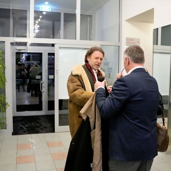 Zwei Männer stehen vor einem Gebäude, einer wird interviewt und hält eine Jacke. Der Interviewer hält ein Mikrofon. Hinter ihnen ist eine offene Glastür mit Leuten im Inneren.