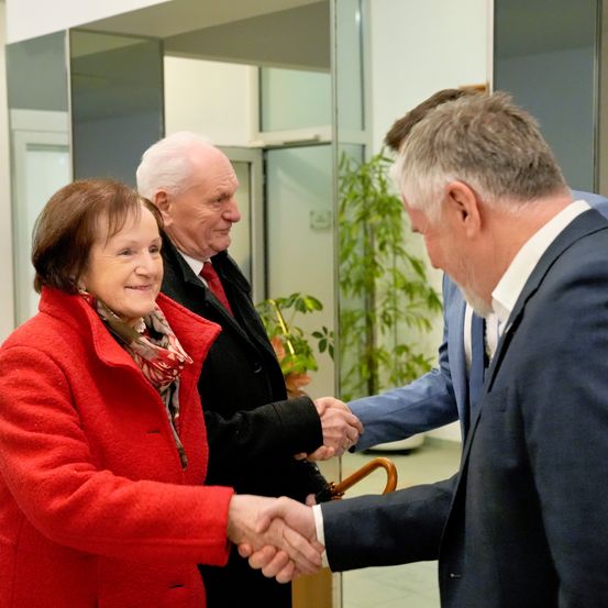 Drei Personen in formeller Kleidung befinden sich in einem Innenraum. Eine Frau in einem roten Mantel schüttelt die Hand eines Mannes in einem blauen Anzug. Hinter ihnen schüttelt ein älterer Mann mit roter Krawatte ebenfalls die Hand des Mannes im blauen Anzug.