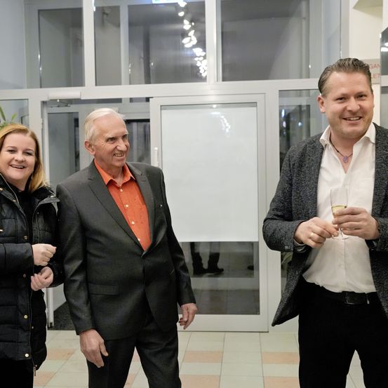 Drei Personen stehen lächelnd vor einer Glastür. Der Mann links trägt einen schwarzen Anzug mit einem orangefarbenen Hemd. Der Mann in der Mitte hat graues Haar und trägt einen dunklen Anzug. Der Mann rechts trägt einen grauen Mantel und hält ein Weinglas.