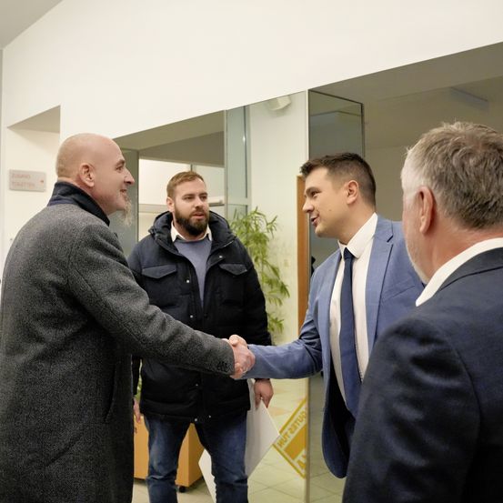 Vier Männer befinden sich in einem Innenbereich, wahrscheinlich einem Büro, mit einer Glastür. Der Mann in der Mitte schüttelt dem Mann auf der linken Seite die Hand. Der Mann auf der rechten Seite schaut sie an. Der Mann auf der linken Seite hält ein Papier. Dahinter steht eine Topfpflanze.