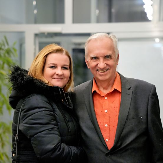 Ein Mann und eine Frau stehen eng beieinander, lächeln und posieren für ein Foto. Die Frau trägt einen schwarzen Mantel und der Mann einen schwarzen Anzug mit einem orangefarbenen Hemd. Sie stehen vor einer Glaswand.