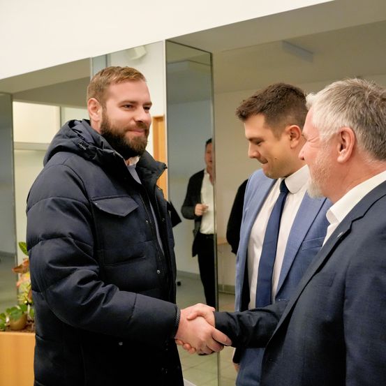 Drei Männer schütteln sich die Hände in einem Raum. Ein Mann trägt eine schwarze Jacke und ein anderer einen blauen Anzug. Ein dritter Mann in einem weißen Hemd steht hinter ihnen.