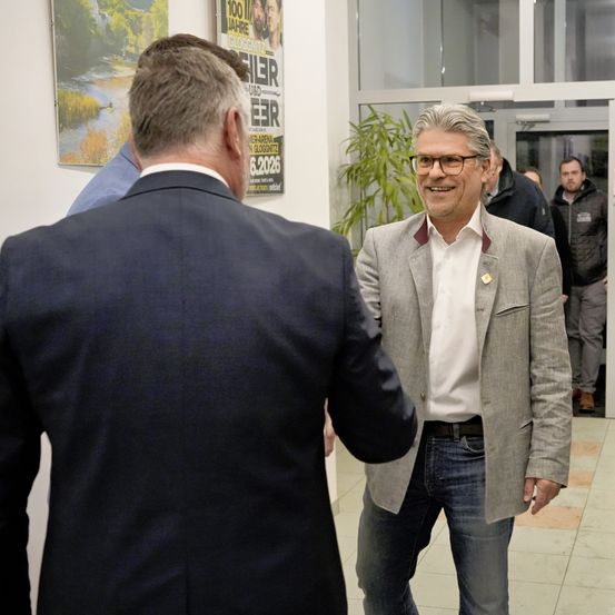 Ein Mann in einem Anzug lächelt, während er durch einen Flur geht und einem anderen Mann im Anzug und einem Mann mit einem Ausweis gegenübersteht.