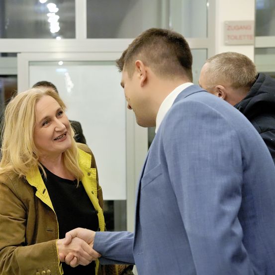 Eine blonde Frau lächelt, während sie einem Mann in einem blauen Anzug die Hand schüttelt. Hinter ihnen steht ein Schild mit der Aufschrift 'Zugang Toilette.' Ein anderer Mann in einem dunklen Mantel schaut zu.