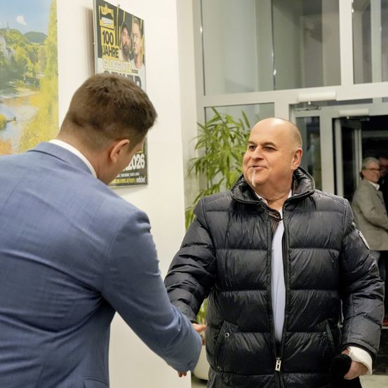 Zwei Männer schütteln sich in einem Foyer die Hände. Einer trägt eine dunkle Jacke, der andere einen blauen Anzug. Es gibt Poster an der Wand und einen Topf mit Pflanzen.