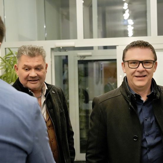 Zwei Männer lächeln sich in einem Bürogebäudeflur an. Beide tragen dunkle Jacken, einer mit Brille. Hinter ihnen befindet sich eine Glastür und eine Pflanze.