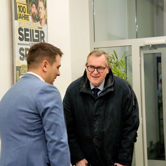 Zwei Männer stehen in einem Innenraum. Ein Mann trägt eine Brille und lächelt. Der andere Mann trägt einen blauen Anzug. Ein Poster ist hinter ihnen.