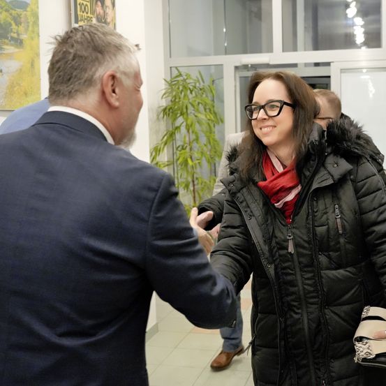 Ein Mann in einem Anzug gibt einer lächelnden Frau in einem schwarzen Wintermantel die Hand, sie stehen in einem Flur mit Topfpflanzen und gerahmten Postern.