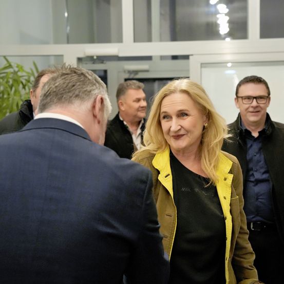 Vier Erwachsene, zwei Männer und zwei Frauen, sind drinnen. Die Frau vor ihnen trägt eine gelbe Jacke und lächelt. Der Mann rechts von ihr trägt eine Brille. Hinter ihnen sind auch ein weiterer Mann und eine Frau anwesend. Der Hintergrund besteht aus Pflanzen und einer Glaswand.