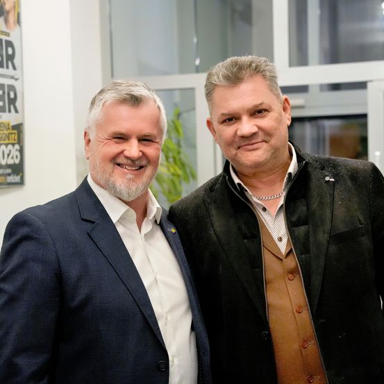 Zwei Männer stehen eng beieinander und lächeln für ein Foto. Der Mann auf der linken Seite trägt einen blauen Anzug. Der Mann auf der rechten Seite trägt einen schwarzen Mantel mit einer braunen Weste. Sie stehen vor einem Glasfenster.