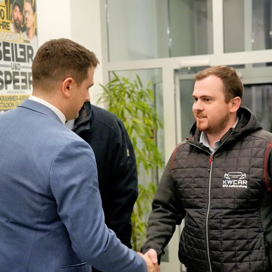 Zwei Männer schütteln sich die Hände in einer Innenraumszene, wahrscheinlich in einem Büro. Der Mann auf der linken Seite trägt einen blauen Anzug, während der Mann auf der rechten Seite eine schwarze Jacke mit einem weißen Logo trägt. Ein Topf mit Pflanzen und eine Glastür sind im Hintergrund zu sehen.