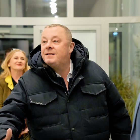 Ein Mann in einem schwarzen Mantel mit Kapuze lächelt und posiert für ein Foto. Hinter ihm blickt eine Frau in einem gelben Shirt zur Seite. Sie stehen in der Nähe einer Glastür mit Lichtreflexionen.