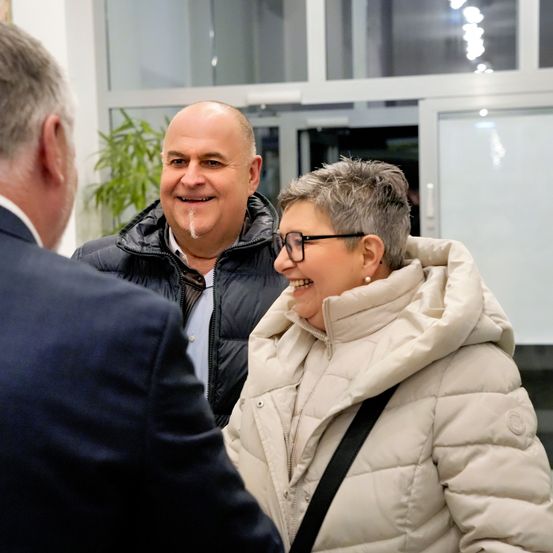 Drei Menschen stehen drinnen, zwei Männer und eine Frau. Der Mann auf der linken Seite trägt einen Anzug und spricht mit dem anderen Mann, der lächelt. Die Frau lacht und trägt eine Brille und eine flauschige Jacke.