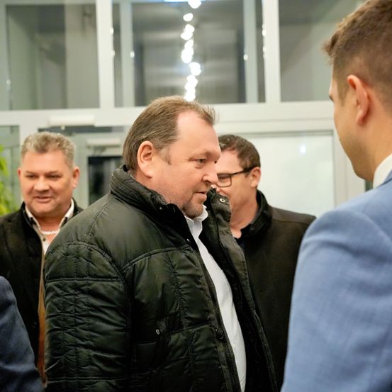 Eine Gruppe von Männern in Winterkleidung steht in einem Raum, einer von ihnen unterhält sich mit einem anderen Mann in einem blauen Anzug.