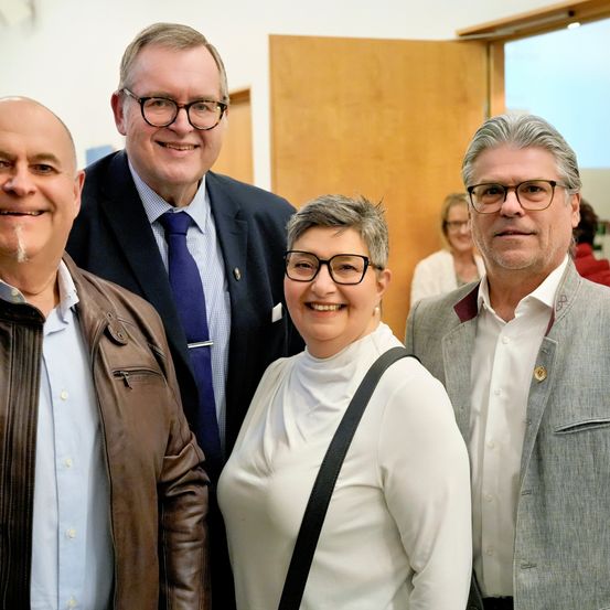 Vier Menschen stehen eng beieinander und lächeln für ein Foto. Der Mann auf der linken Seite trägt eine braune Jacke, während die anderen in formeller Kleidung sind. Hinter ihnen sieht eine Frau zu.