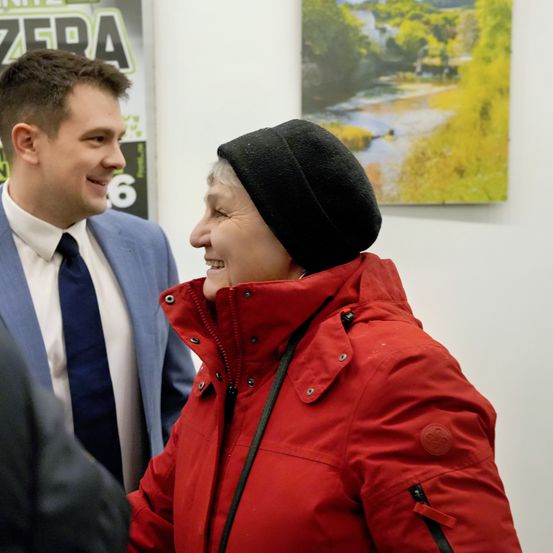 Ein Mann in Anzug und Krawatte lächelt und unterhält sich mit einer älteren Frau in einem roten Wintermantel und schwarzer Mütze. Sie stehen vor einer weißen Wand.