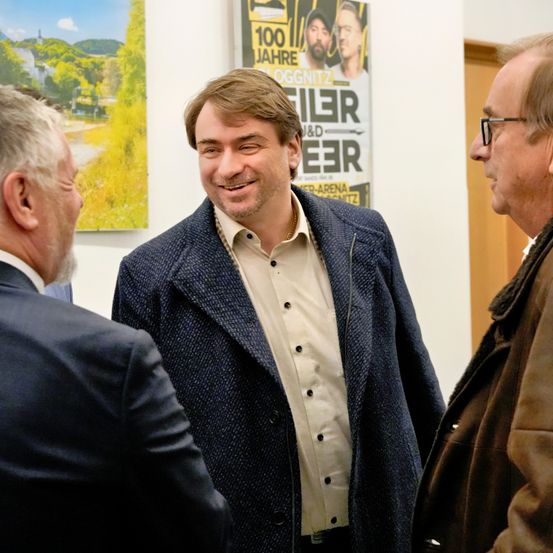 Ein Mann in einem blauen Mantel lächelt und unterhält sich mit zwei Männern in Anzügen. Hinter ihnen befindet sich ein Plakat mit den Worten '100 Jahre OGGNITZ' und einem Bild von zwei Männern.