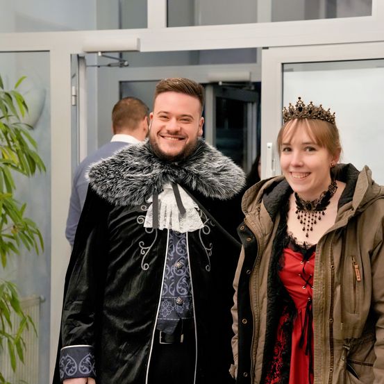 Ein Mann und eine Frau in mittelalterlichen Kostümen lächeln und stehen neben einer Glastür. Hinter ihnen steht ein weiterer Mann in einem Anzug.