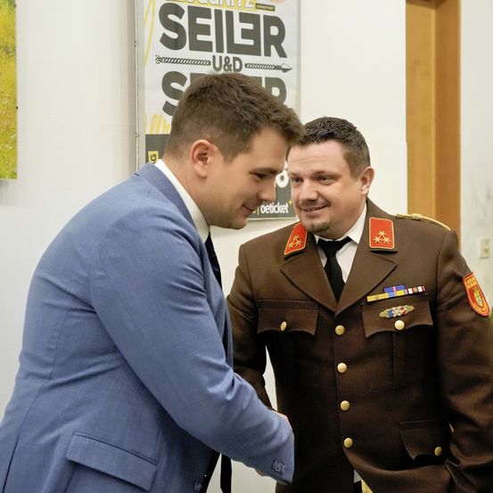 Zwei Männer stehen sich gegenüber. Der Mann im blauen Anzug schüttelt dem Mann in der Militäruniform die Hand, beide lächeln. Hinter ihnen wirbt ein Poster für Seiler.