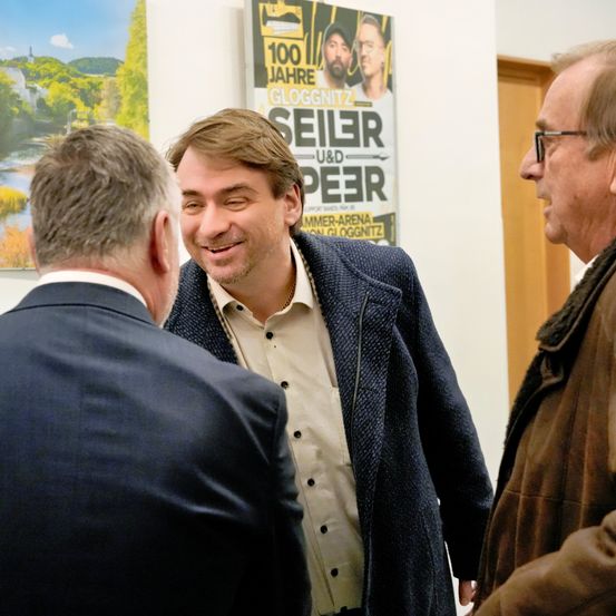 Drei Männer stehen in einem Flur. Ein Mann trägt eine Brille und einen braunen Mantel, während ein anderer Mann lächelt und einen blauen Mantel trägt. Ein Poster mit Text steht hinter ihnen.