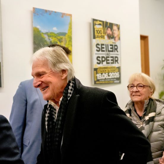 Ein Mann mit weißen Haaren lächelt und schaut in die Kamera. Hinter ihm stehen zwei Personen. An der Wand hängen zwei Poster. Eines der Poster hat den Text 'SeiLer und Speer' und ein Datum.